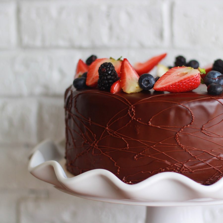 Pastel de chocolate con frutos rojos sobre base blanca, frente a una pared de ladrillo blanco
